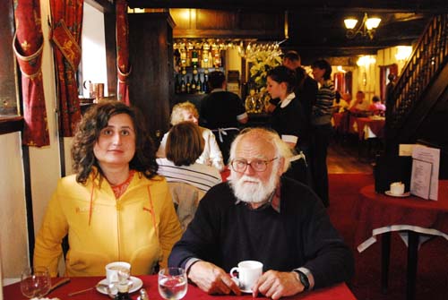 Peter and Maggie are sitting at a table having tea at the famous De Greys cafe. Maggie has shoulder length brown hair and is wearing a yellow jacket. Peter has white hair and a white beard and is wearing a blue jersey. Behind them are the waitresses in their black dresses and white aprons.